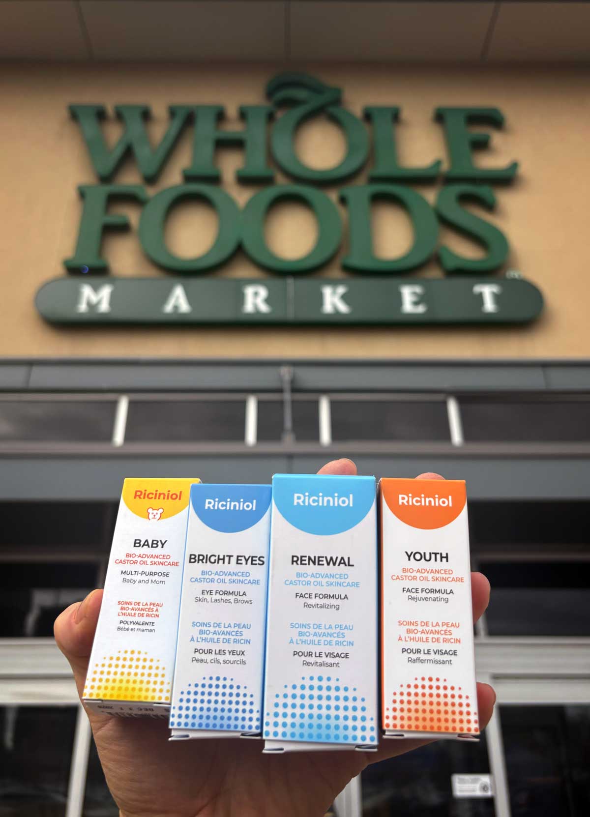 Four Riciniol product boxes held up in front of the Whole Foods Market sign